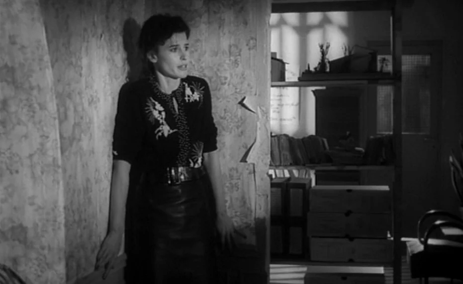 Vivement Dimanche (1983) movie still: medium — A dimly lit room features a patterned wallpapered wall and shelves with stacked boxes. A woman leans…
