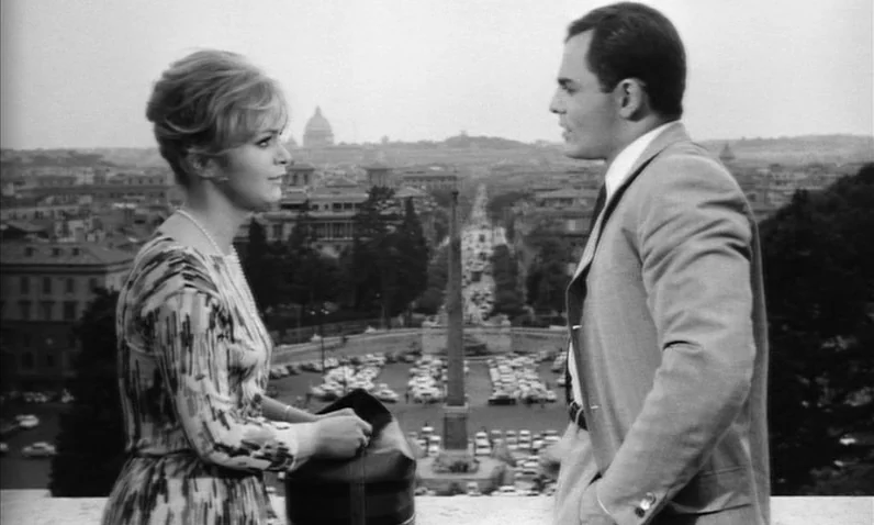 The Girl Who Knew Too Much (1963) movie still: medium — A woman and a man stand on a stone balcony overlooking a Roman square. An obelisk stands in…
