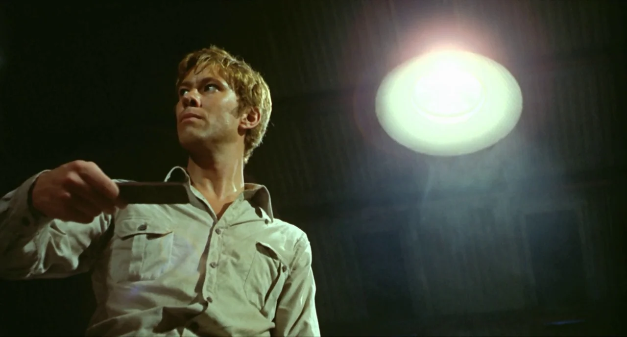 Wake In Fright (1971) movie still: medium — An industrial interior features dim, shadowy rafters and a single, starkly lit area. A man stands under…