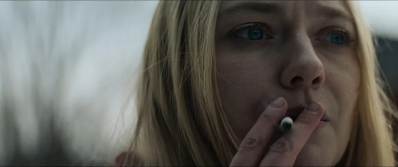 Vicious (2025) movie still: closeup — An overcast outdoor setting with blurred background elements. A woman with blonde hair holds a cigarette to…