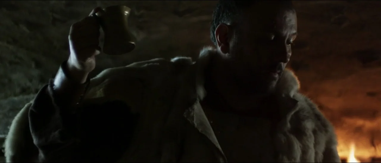 The Penitent Thief (2021) movie still: medium — A dark, textured stone enclosure is illuminated by low firelight from the bottom right. A bearded…