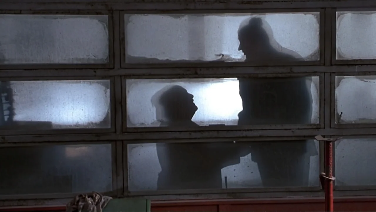 Matilda (1996) movie still: medium — A workshop interior featuring a wall of rectangular frosted glass windows and red support beams. Two figures…