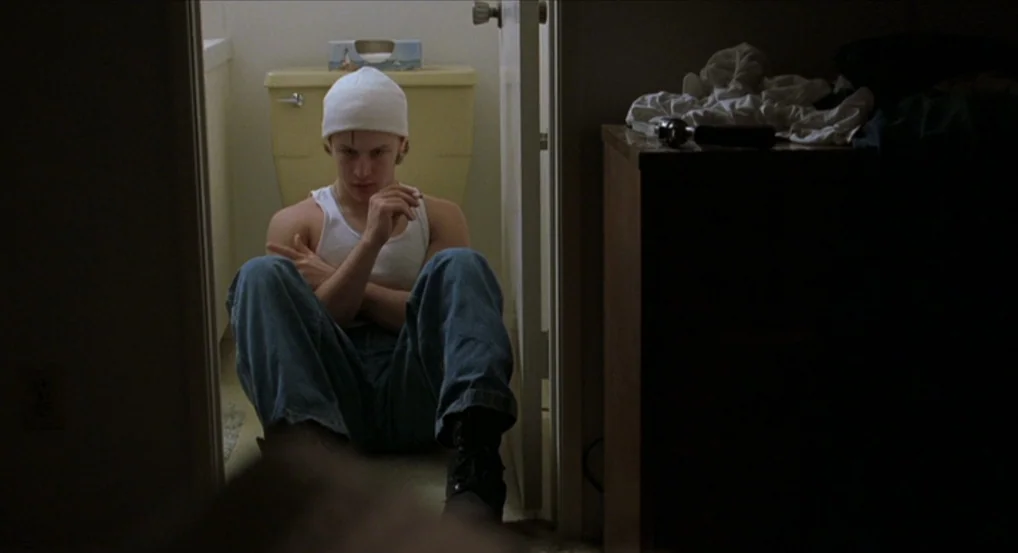 Brick (2005) movie still: medium — A man sits on the floor inside a cramped, dimly lit residential bathroom. He rests against the wall near the…