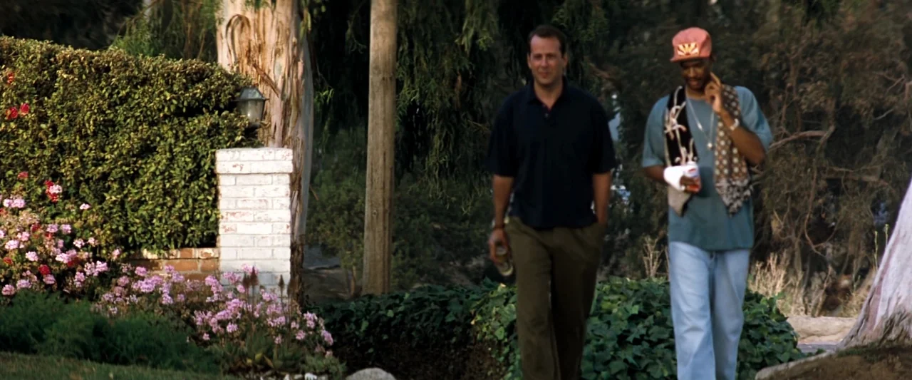 The Last Boy Scout (1991) movie still: fullbody — A residential sidewalk bordered by a hedge and a tree-lined background. Two men walk side by side…