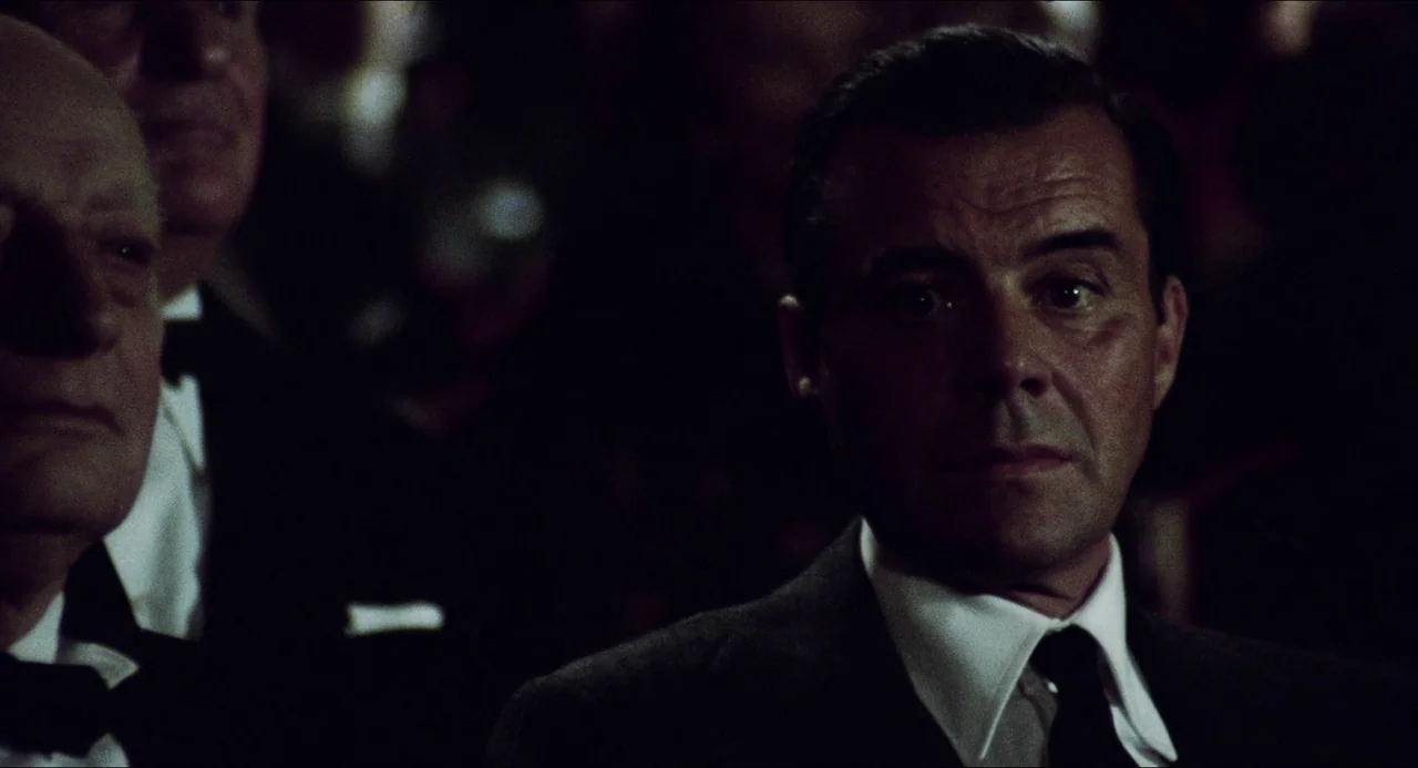 The Night Porter (1974) movie still: medium — Two men in formal attire sit in a dark room with rows of seats. A man in the foreground looks forward…
