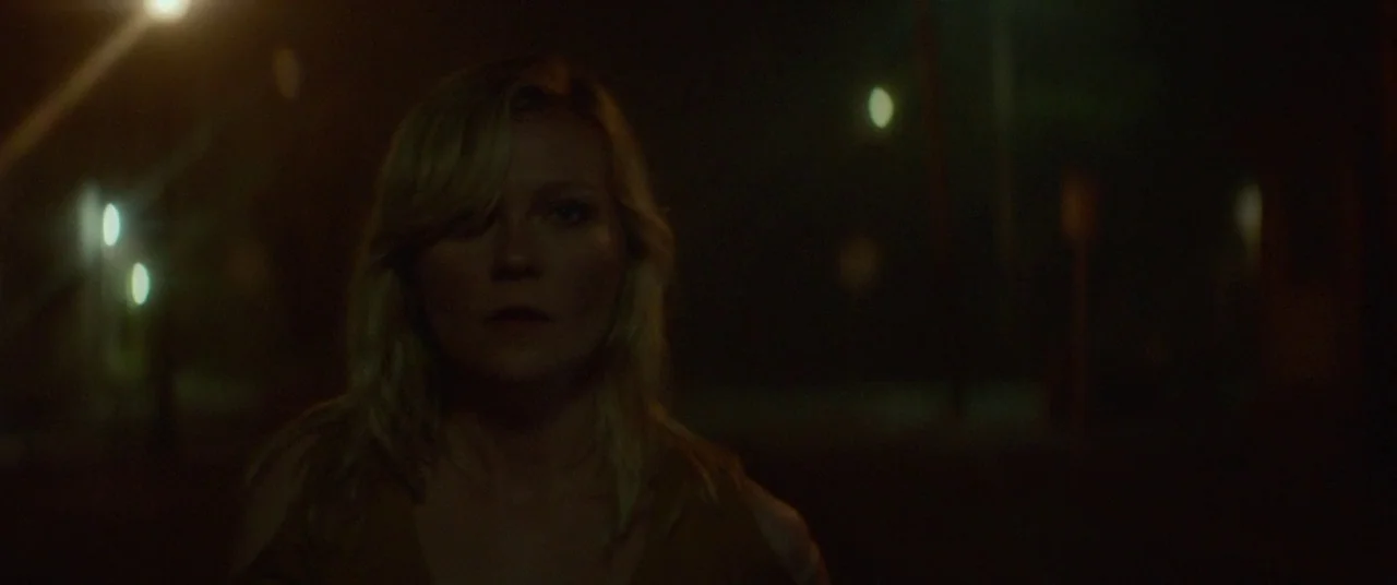 Woodshock (2017) movie still: medium — A dark night setting outdoors with bokeh lights visible from distant streetlamps. A woman walks forward in…