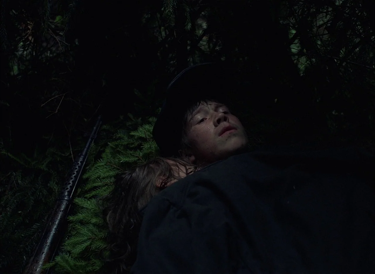 Come and See (1985) movie still: closeup — Dense, dark pine needles surround a young boy lying on the damp ground. The boy rests his head against an…