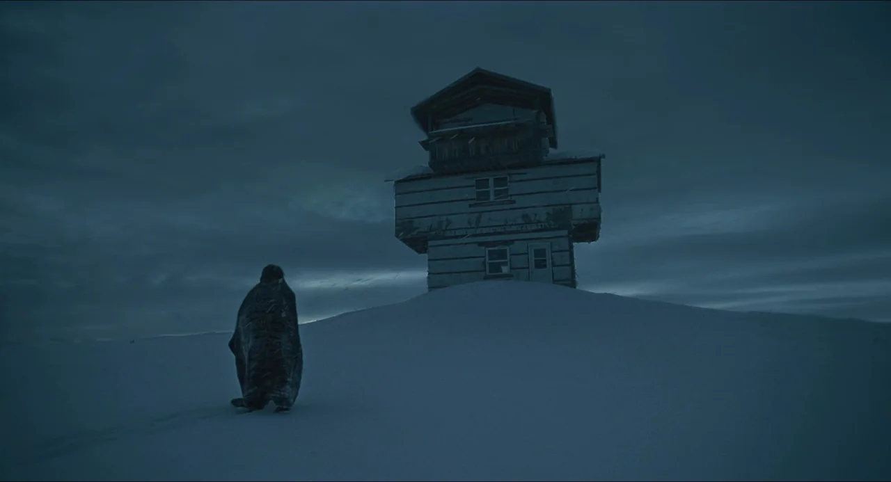 The Lodge (2019) movie still: wide — An isolated wooden structure stands atop a snow-covered hill under a dark sky. A person draped in a heavy dark…