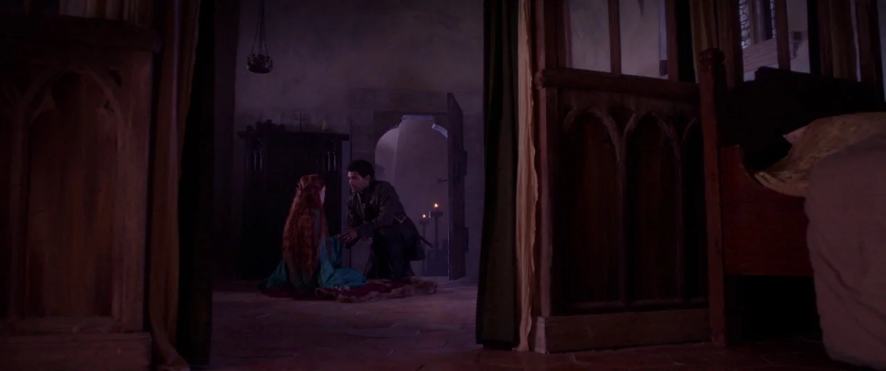 Ophelia (2018) movie still: wide — A stone chamber features dark wooden screens and a fur rug on the floor. A man kneels on the floor holding a…