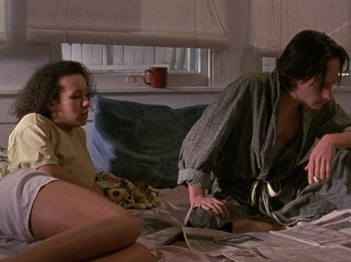 Slacker (1991) movie still: medium — A light-filled room with window blinds behind a bed covered in newspapers. A man in a robe reads a paper while…