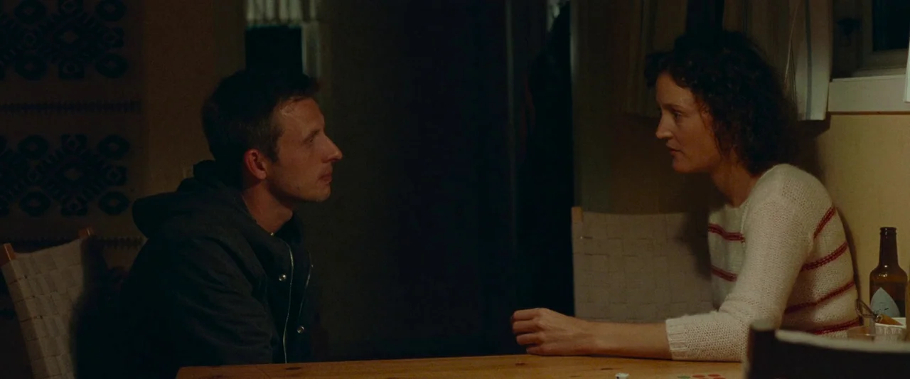 Bergman Island (2021) movie still: medium — A dimly lit room with a wooden table where two people sit opposite each other. The space features woven…