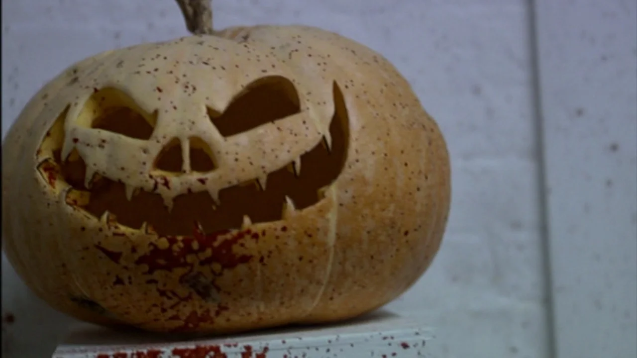 Murder Party movie still: closeup — A pale, carved pumpkin sits on a white rectangular base against a white wall. The pumpkin surface is heavily…