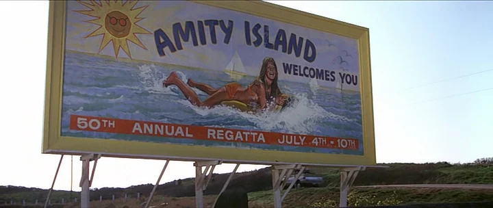 Jaws movie still: fullbody — A large painted wooden billboard stands on a rural roadside against a clear sky. The sign advertises Amity Island and…