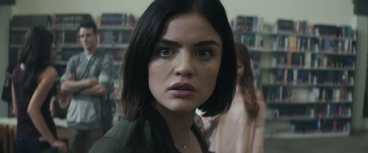 Truth or Dare (2018) movie still: closeup — A quiet room filled with tall metal shelving units packed with various hardcover books. A young woman in…