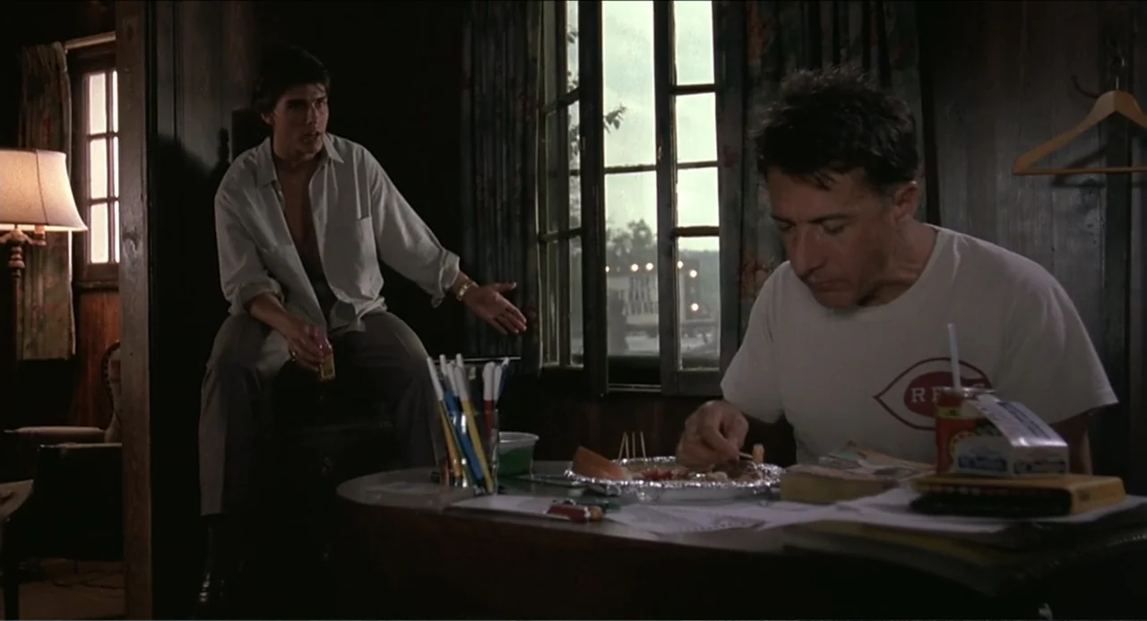 Rain Man (1988) movie still: medium — A wood-paneled room contains a table covered in papers, pens, and food. One man sits at the table eating while…
