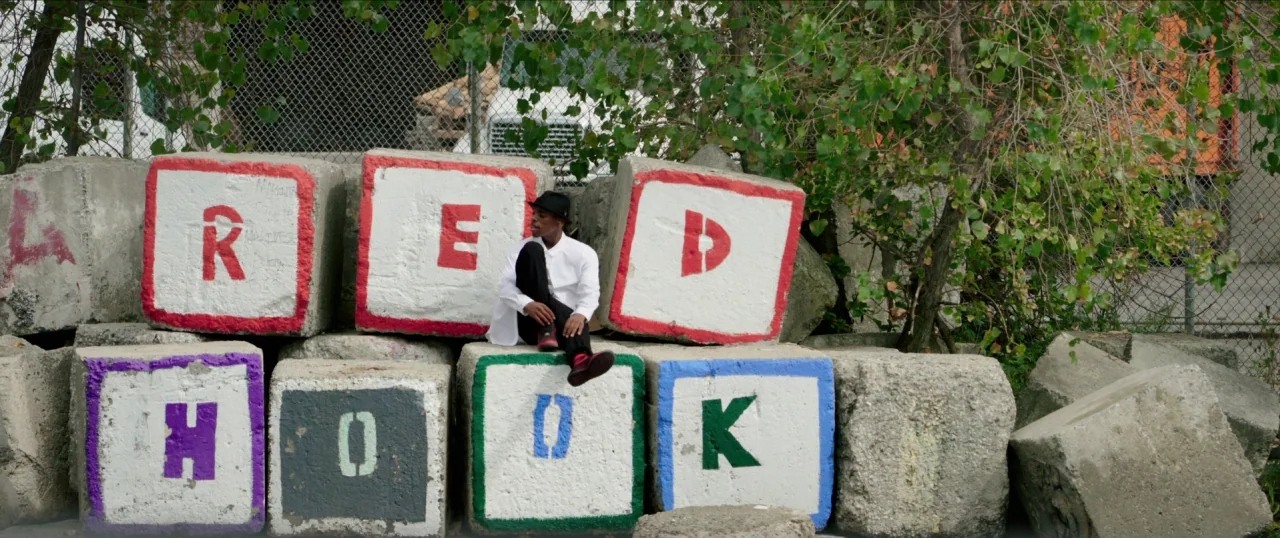 Da Sweet Blood of Jesus (2014) movie still: wide — A pile of large concrete blocks with painted letters spelling Red Hook sits outdoors. A young man…