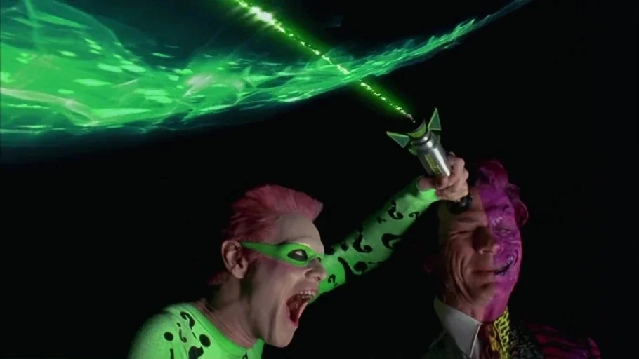 Batman Forever (1995) movie still: medium — Two men stand before a dark, featureless backdrop with a bright green energy effect. The Riddler reaches…