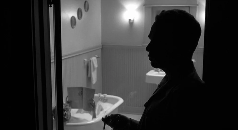 The Man Who Wasn’t There (2001) movie still: medium — A vintage tiled bathroom with a clawfoot tub and wall-mounted light fixture. A silhouette of a…