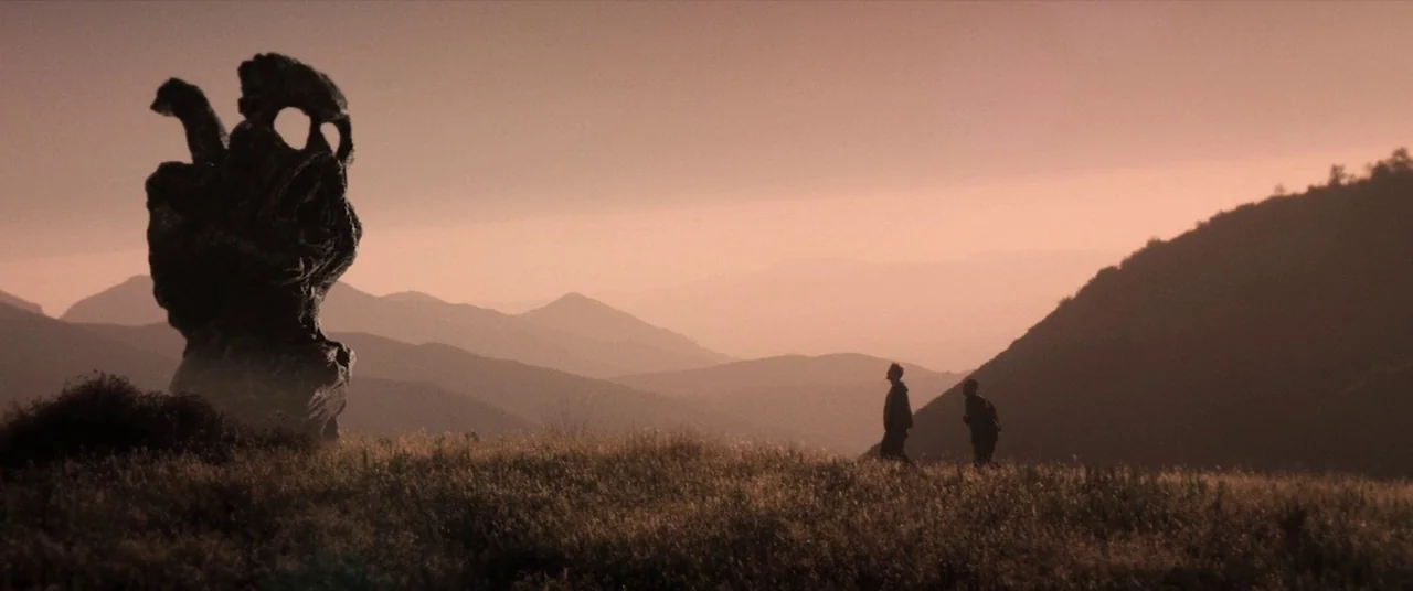 The Endless (2017) movie still: wide — A grassy hill at sunset features a large, dark, abstract stone sculpture. Two men walk across the dry grass…