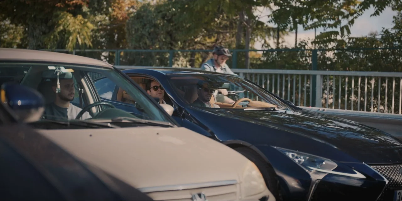 Kill Me If You Dare (2019) movie still: medium — A multi-lane road holds multiple cars stopped in daytime traffic. Two men sit in separate cars…