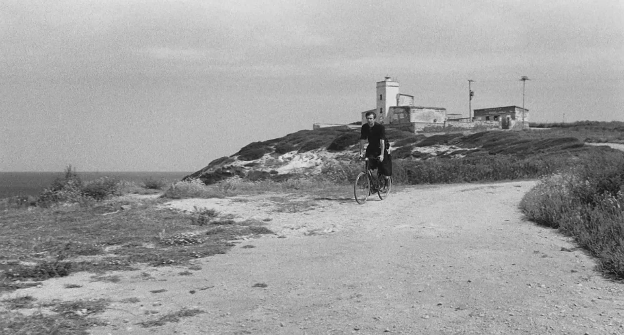 The Legend Of Kaspar Hauser (2012) movie still: wide — A man rides a bicycle on a dusty path near a desolate coastline. In the background, a white…