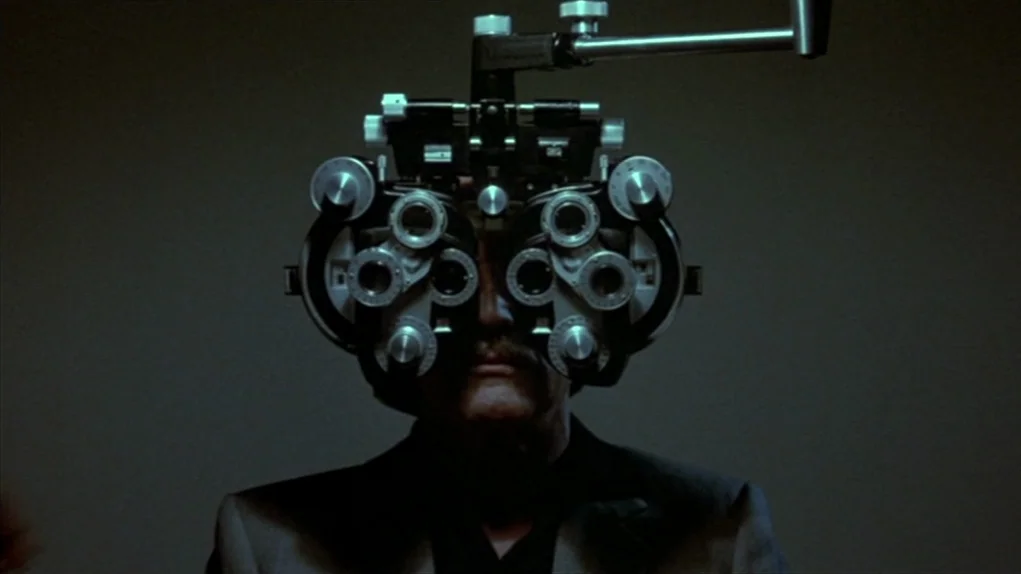 The Colour of Money (1986) movie still: closeup — A man is positioned in a dark room with a complex optometric testing device. He looks forward…
