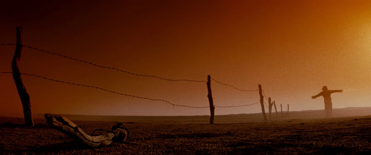 Razorback (1984) movie still: wide — A vast, empty field is intersected by a long, weathered barbed wire fence. A single scarecrow stands near the…