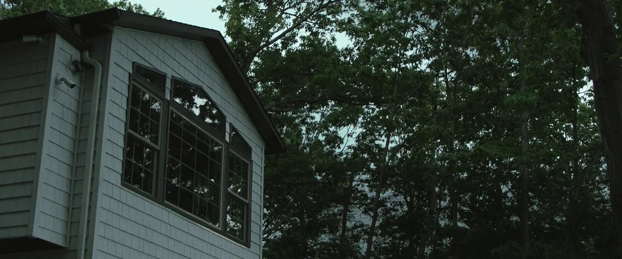 The Night House (2020) movie still: wide — A light-gray suburban house exterior sits next to a thick stand of mature trees. The overcast sky creates…