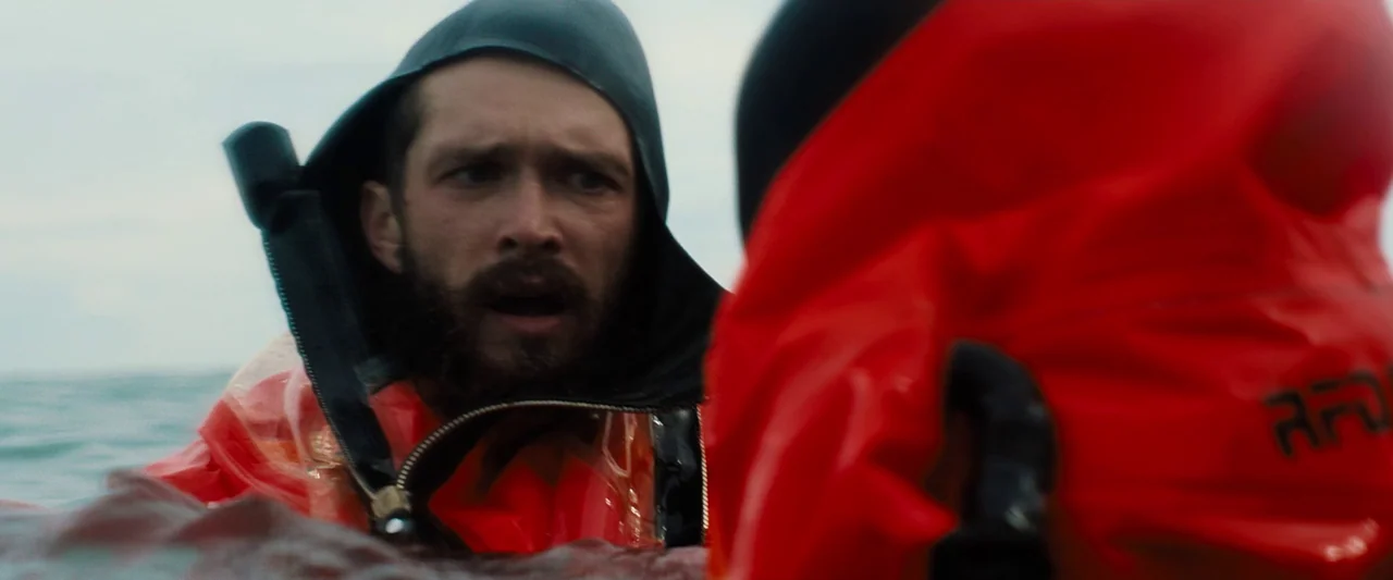 Black Sea (2014) movie still: closeup — An overcast sky meets a choppy sea surface with limited visibility. A man with a dark beard wearing a bright…