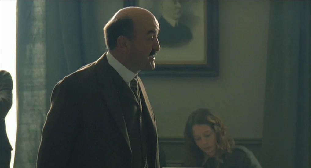 The Wind That Shakes the Barley (2006) movie still: medium — An interior office space with muted walls and a framed portrait on the wall. A man in a…