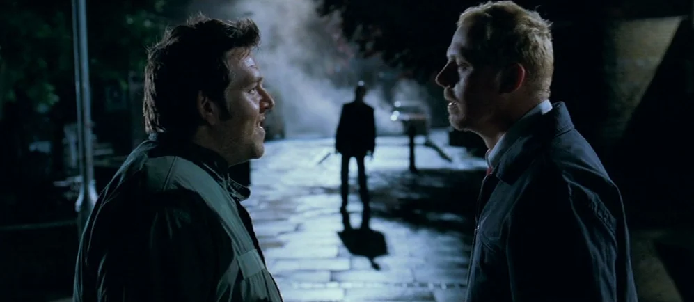 Shaun Of The Dead (2004) movie still: medium — A dark, wet cobblestone street at night is illuminated by a single overhead light source. Two men…