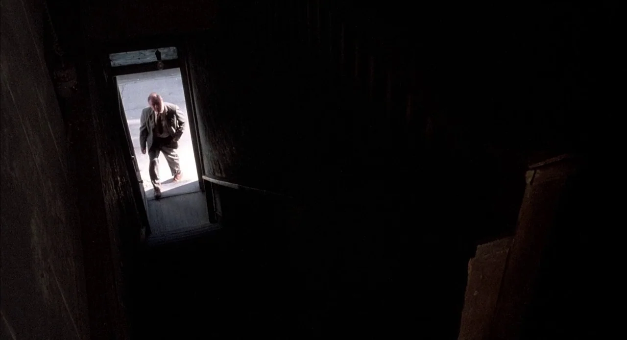Mississippi Burning (1988) movie still: wide — A dark, unlit stairwell leads up to a bright doorway at the top landing. A middle-aged man in a suit…