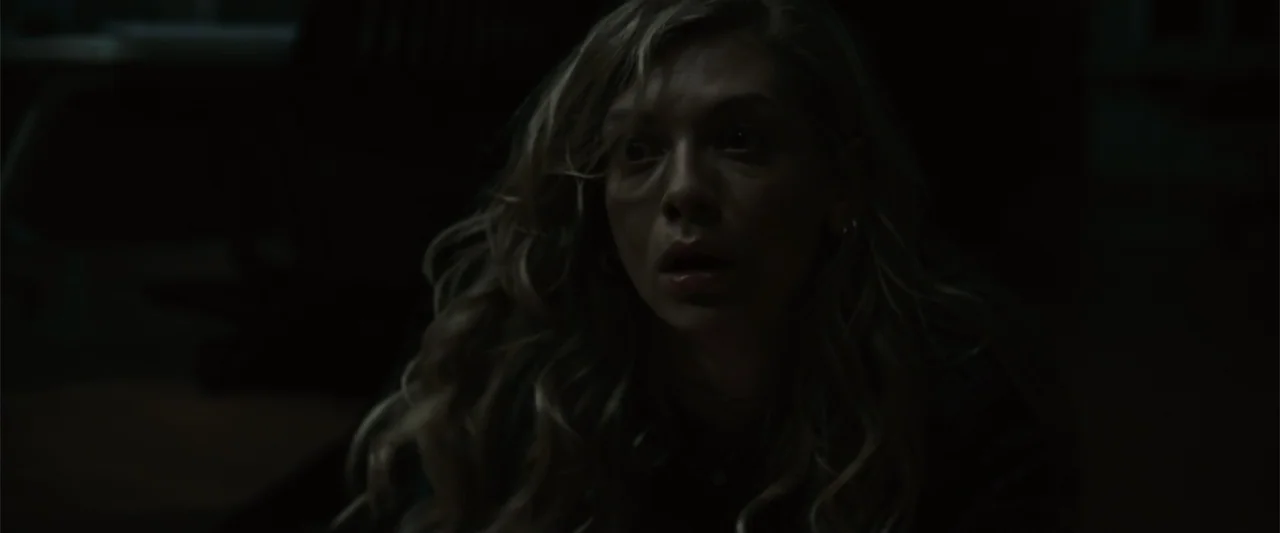 Init!ation (2021) movie still: closeup — A dark, indistinct indoor space is shrouded in deep shadows. A young woman with blonde hair stares…
