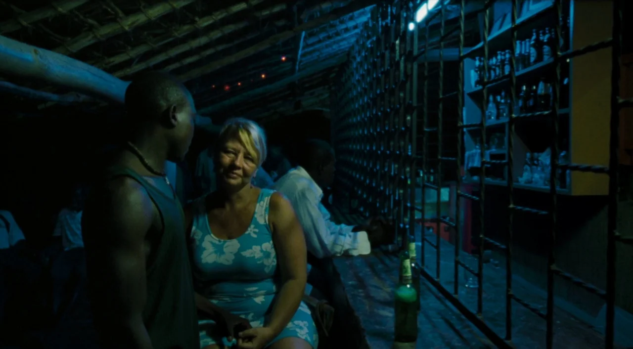 Paradise: Love (2012) movie still: medium — A dimly lit bar with a bamboo ceiling and metal barred shelves holding bottles. A woman sits at the bar…