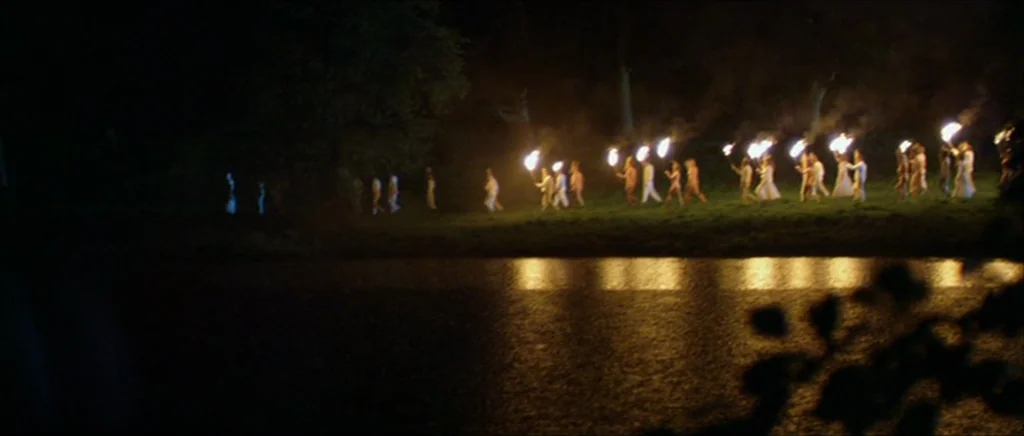 Kill List movie still: wide — A group of people wearing light clothing walks in a line by a lake at night. They carry torches that cast light across…
