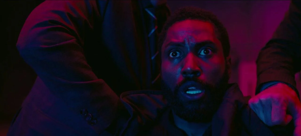 Tenet (2020) movie still: closeup — A dark space lit by sharp red and blue neon light sources. A man with wide eyes looks toward the camera with his…