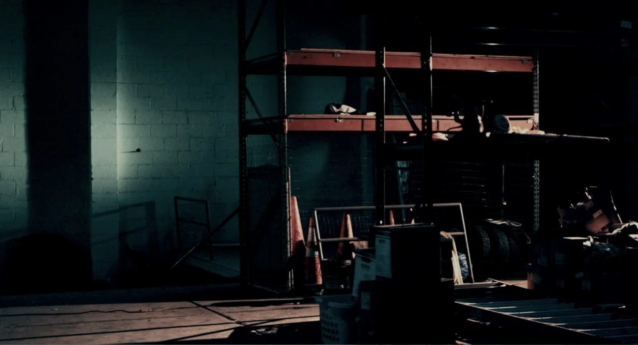 Terrifier (2018) movie still: establishing — A dark, industrial space features metal storage racks against a white concrete block wall. Empty floor…