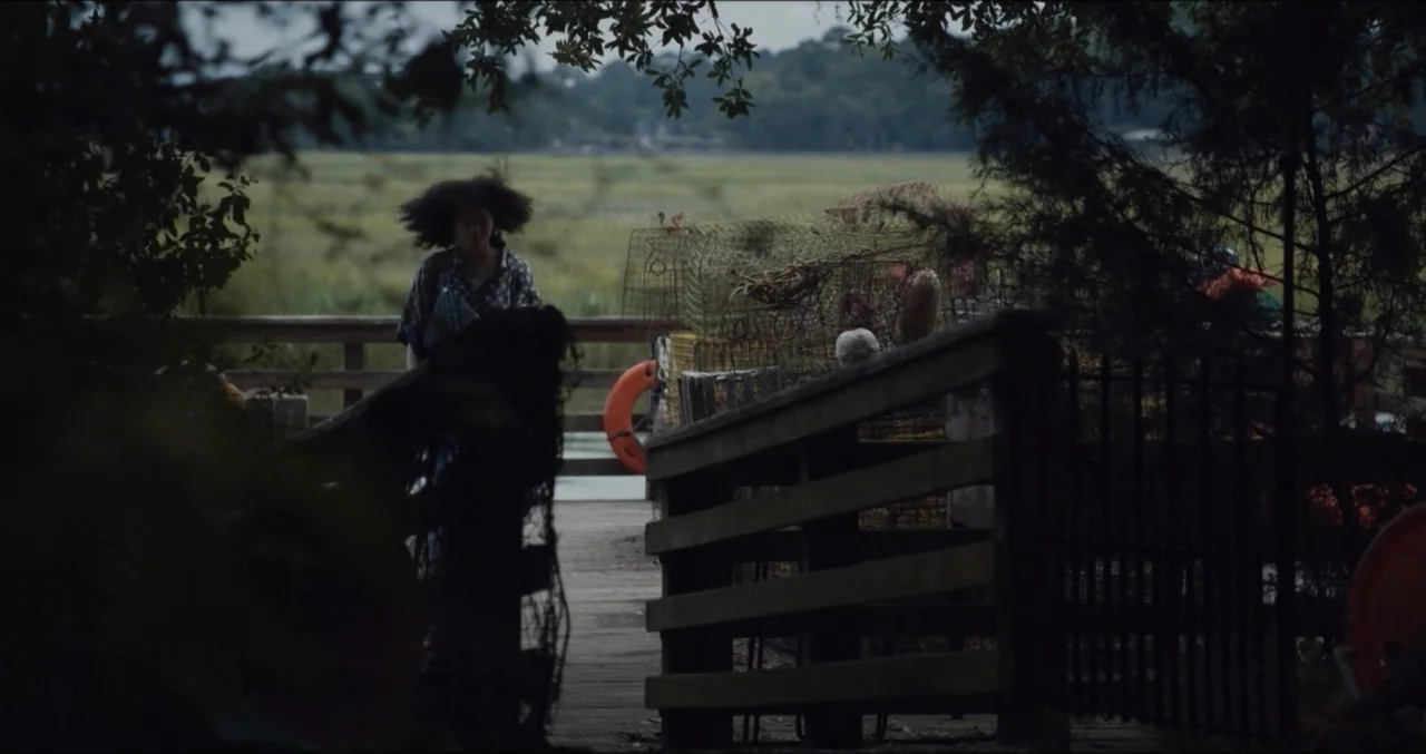 Eye for an Eye (2025) movie still: medium — A wooden pier extends over a marshy landscape under an overcast sky. A woman walks toward the camera…