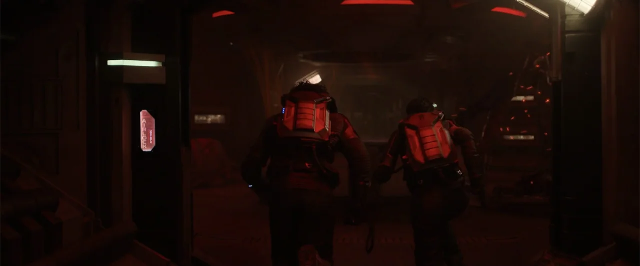DISTANT (2026) movie still: medium — A dimly lit industrial corridor inside a spacecraft features red emergency lighting. Two figures wearing…