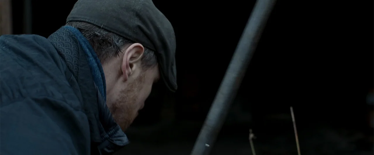 My Son (2021) movie still: closeup — A dark, empty barn interior with a single light source defining the foreground space. A man wearing a flat cap…
