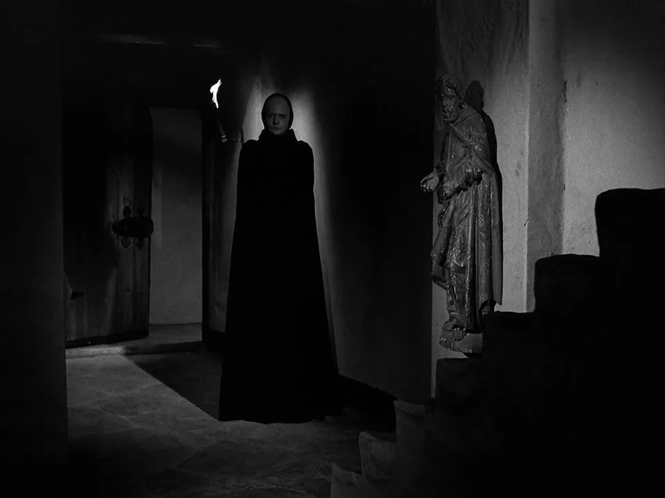 The Seventh Seal (1957) movie still: fullbody — A dark stone corridor features a carved religious statue attached to the wall. A man in a dark…