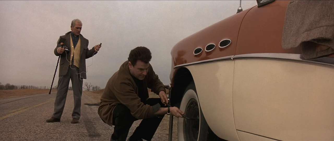 Cobb (1994) movie still: wide — A rural asphalt road stretches past a vintage two-tone car on an overcast day. One man kneels to tighten a lug nut…