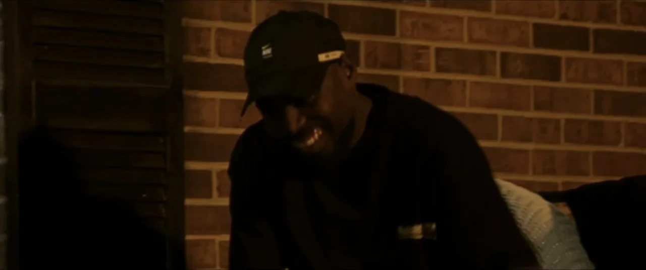 Sermon on the Mount (2019) movie still: medium — A man stands in front of a dark brick wall at night. The man wearing a black cap smiles broadly…