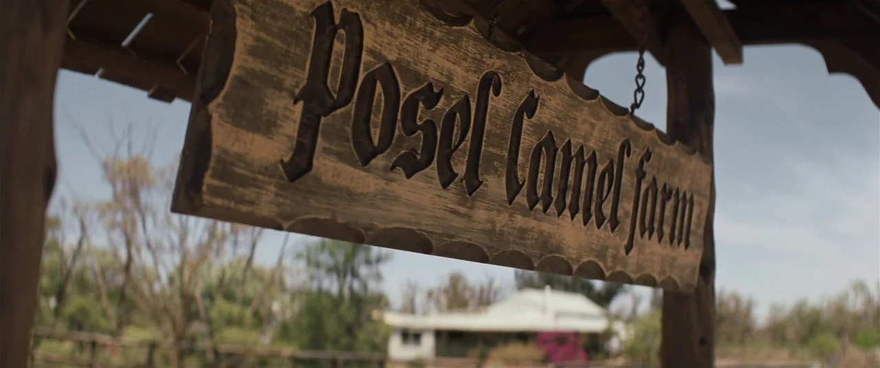 Tracks (2013) movie still: closeup — A wooden sign with carved lettering reading Posel Camel farm hangs under a shelter. The sign is suspended by…