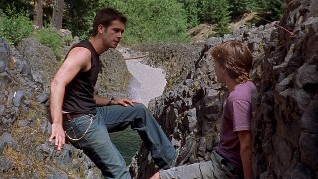 Mean Creek (2004) movie still: medium — A narrow rocky passage features jagged dark stone walls and a small flowing stream. Two people stand and sit…