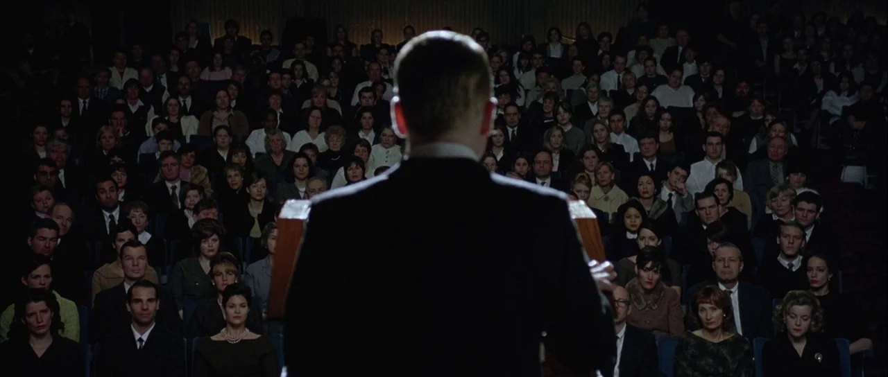 Capote (2005) movie still: medium — A large crowd fills rows of seats inside a dimly lit dark wood auditorium. A man stands at a wooden podium in…