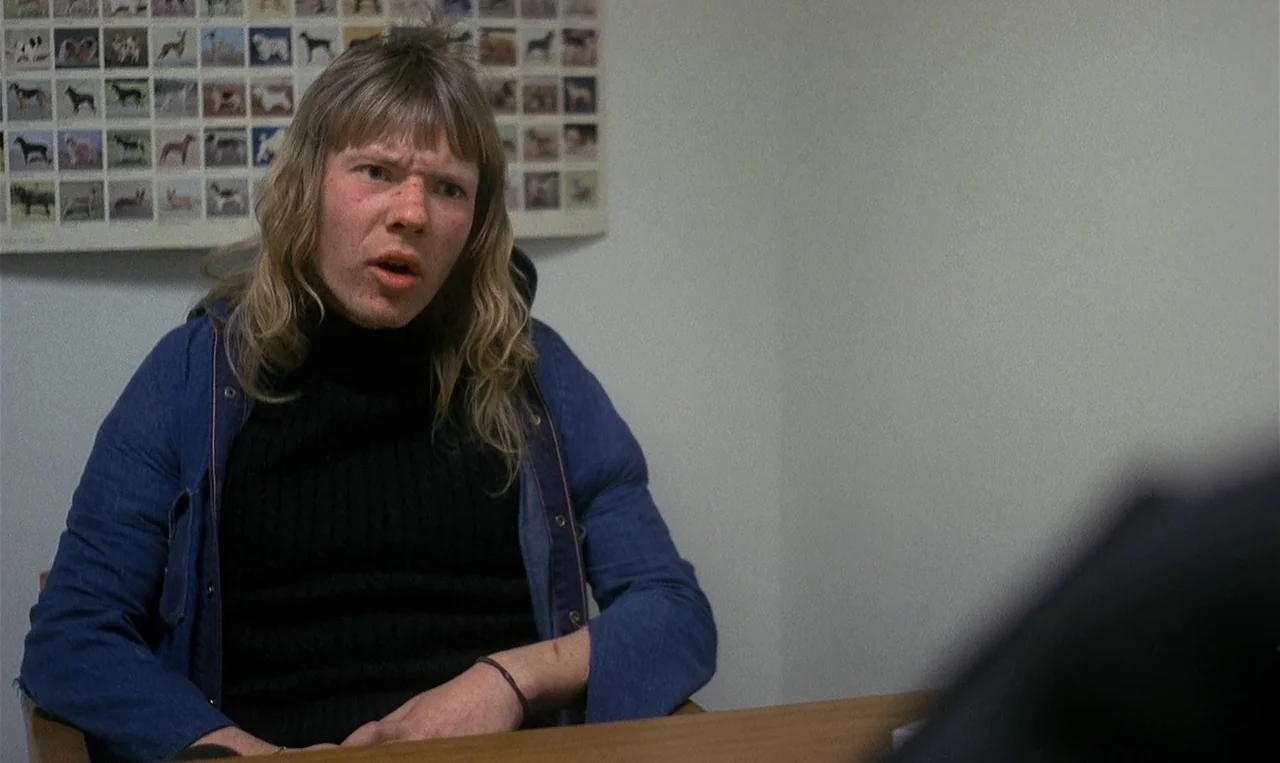 Man on the Roof (1976) movie still: A tense, realistic conversation captured in a sterile office environment. The lighting is unflattering and raw,…