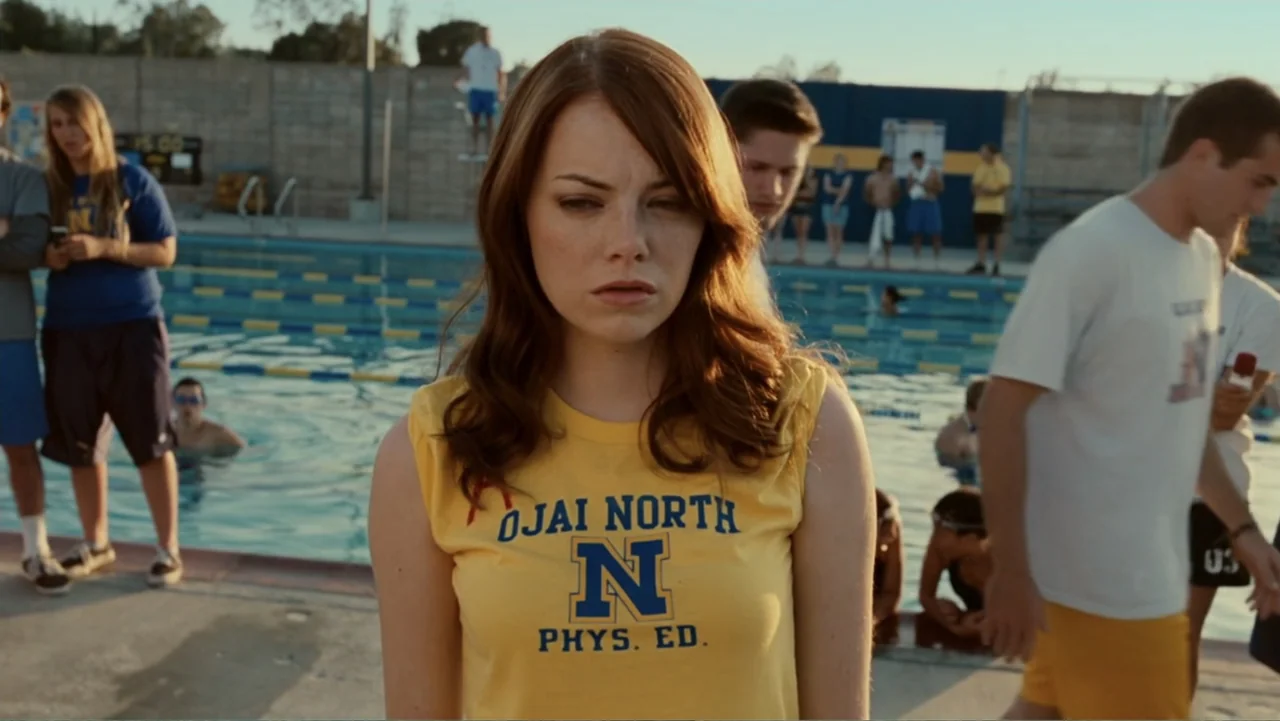 Easy A (2010) movie still: medium — A bustling public swimming pool deck with teenagers milling about in athletic gear. A young woman in a yellow…