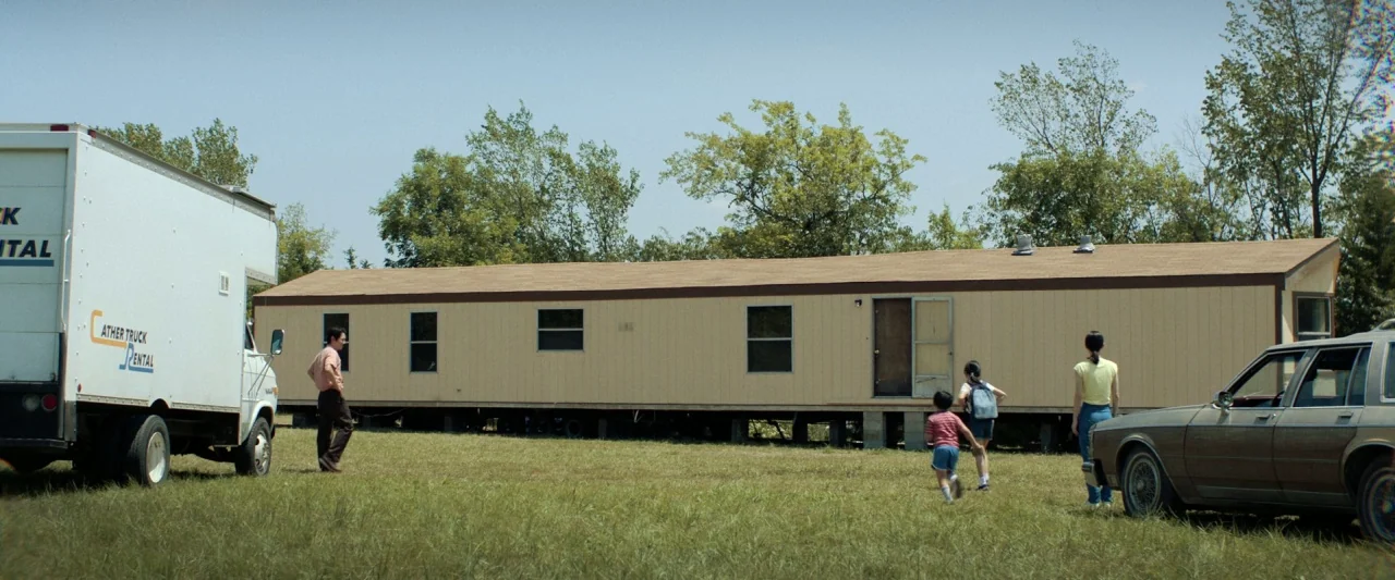 Minari (2020) movie still: wide — A tan mobile home sits on a dry grass lot under a clear blue sky. A man, a woman, and two children walk toward the…