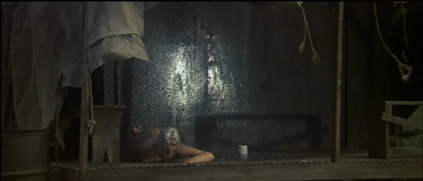 MASH (1970) movie still: wide — A makeshift wooden enclosure stands outdoors with hanging ropes and wet wooden slats. A man lies prone on the…